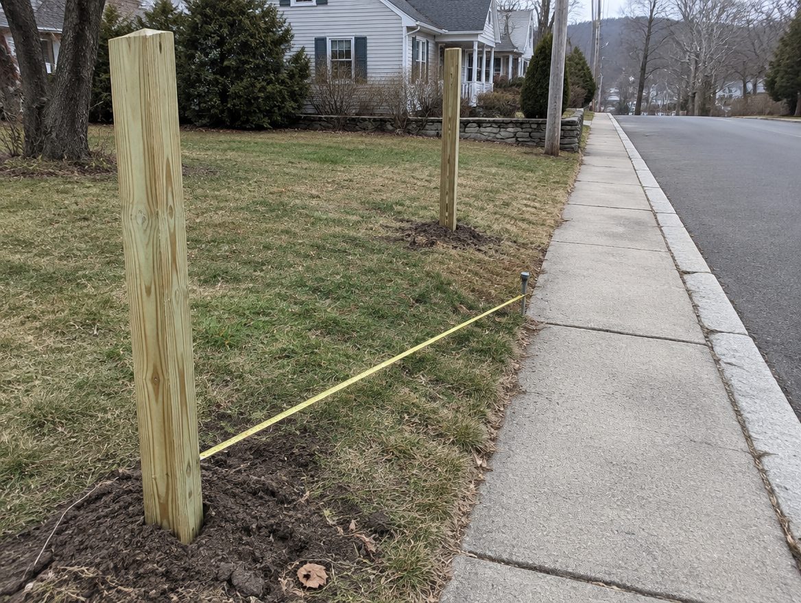 Fence posts set at the front corner of a village lot beside a sidewalk, with a measuring tape across to the property pin
